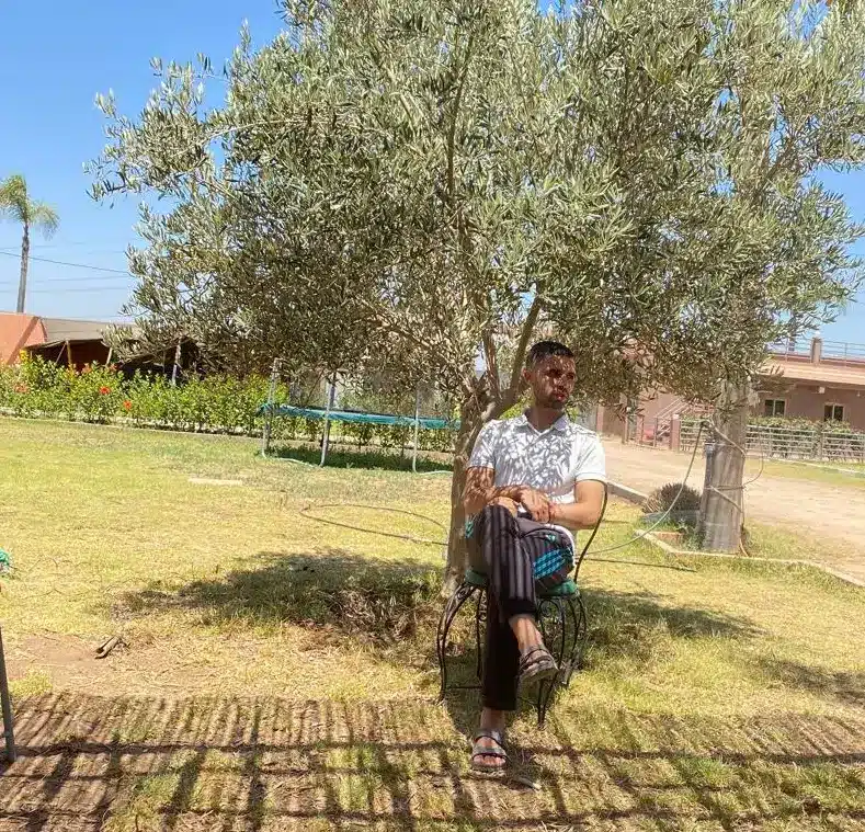 Young Moroccan man sitting peacefully under an olive tree in a sunny rural garden.