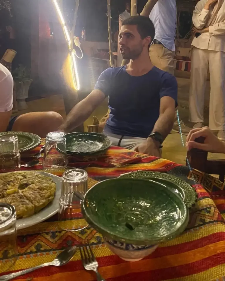 Man enjoying traditional Moroccan dinner outdoors with colorful Amazigh tablecloth, green ceramic bowls, and a local dish under soft lighting.