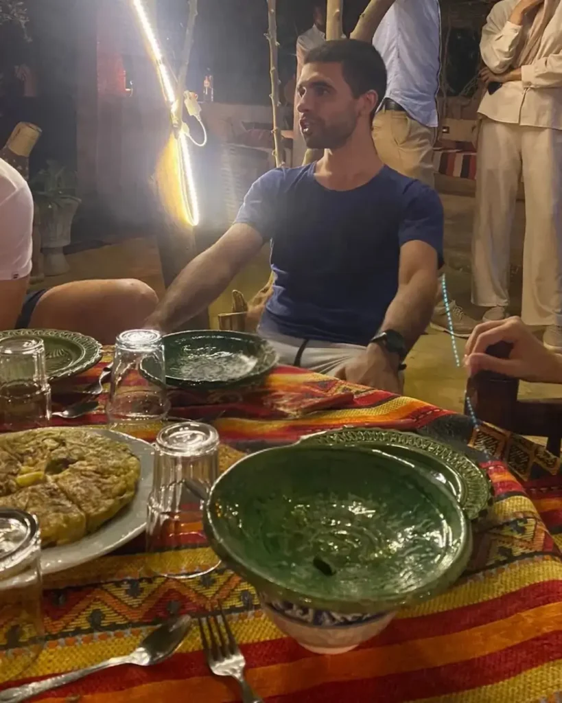 Man enjoying traditional Moroccan dinner outdoors with colorful Amazigh tablecloth, green ceramic bowls, and a local dish under soft lighting.